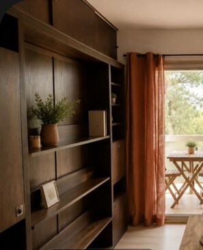 A room with a wooden bookshelf, a small table with a plant, and a view through a window with curtains. - Studio with a view of the ocean (Cavalaire-sur-Mer)