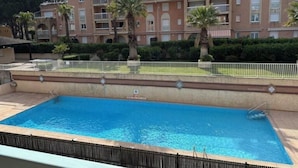 Una piscina con valla perimetral, un edificio con varios balcones y palmeras.