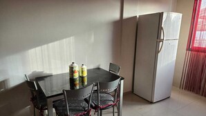 Ruokailutila, jossa on pöytä ja tuolit, jääkaappi ja ikkuna, jossa on sälekaihtimet.