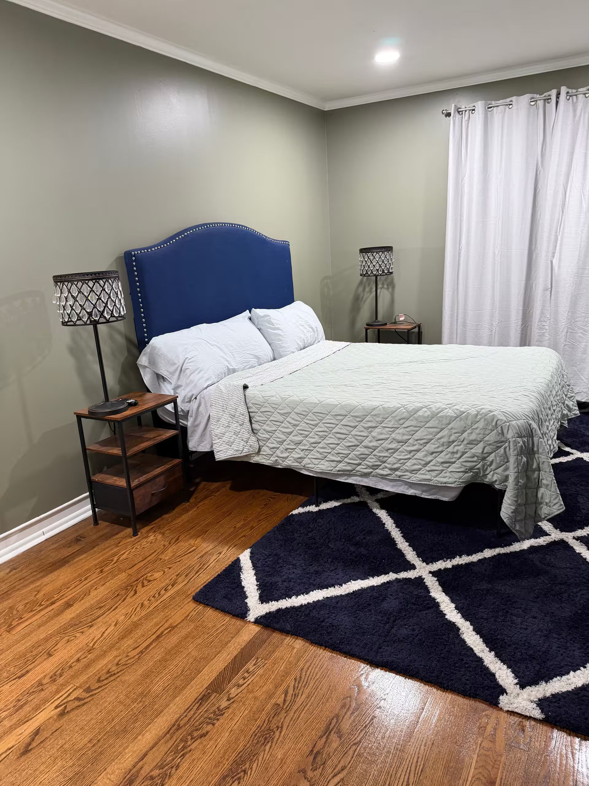 A bedroom with a bed, two bedside lamps, a nightstand, a dark blue rug, and wooden flooring.