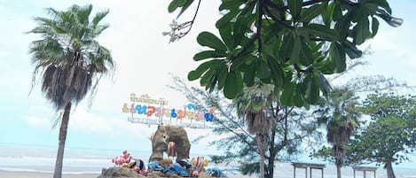 Un paseo marítimo en la playa con palmeras, una escultura de piedra y un cartel.