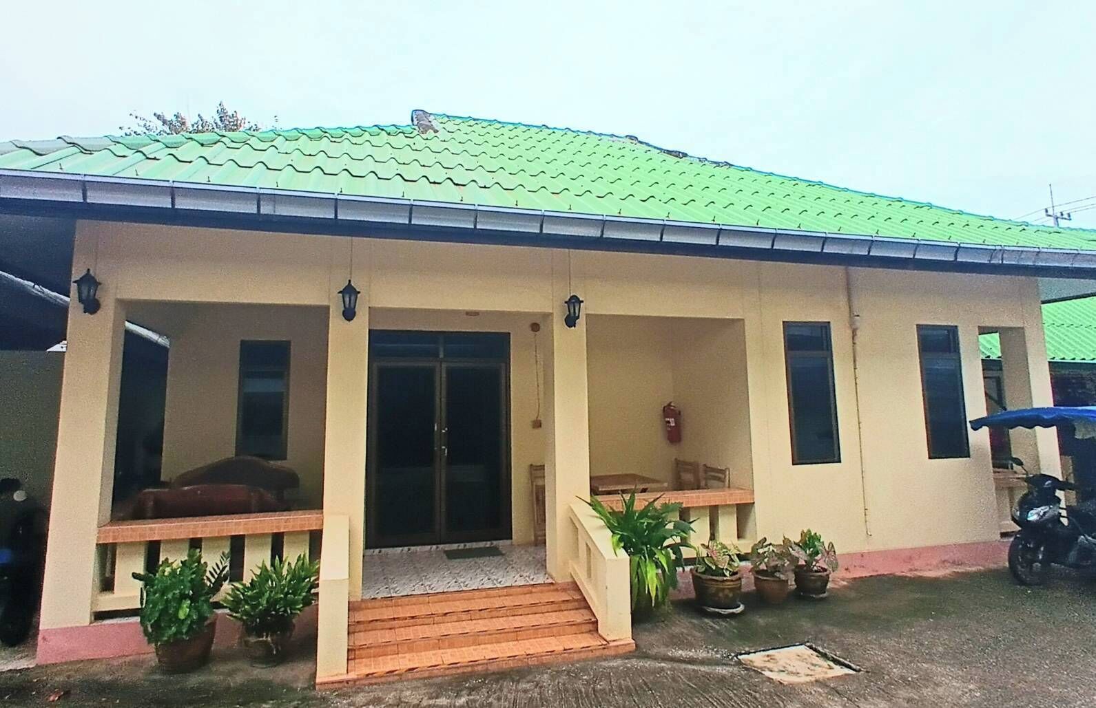 A single-story building with a green roof, a covered porch with potted plants, and a visible fire extinguisher.