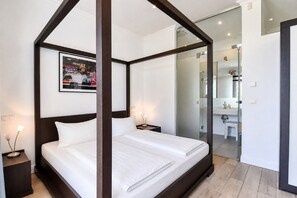 A modern bedroom with a four-poster bed, a bedside table with a lamp, a framed picture on the wall, and a bathroom visible through a glass door. - Vacation Rental at Villa Rex Sellin – Pool, Sauna, and the Sea (Sellin)