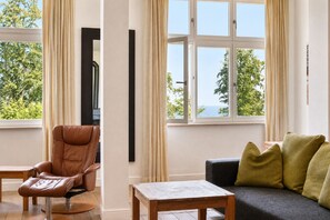 Um quarto com poltrona de massagem, sofá com almofadas, pequena mesa de madeira e janelas amplas com vista para árvores e o mar.