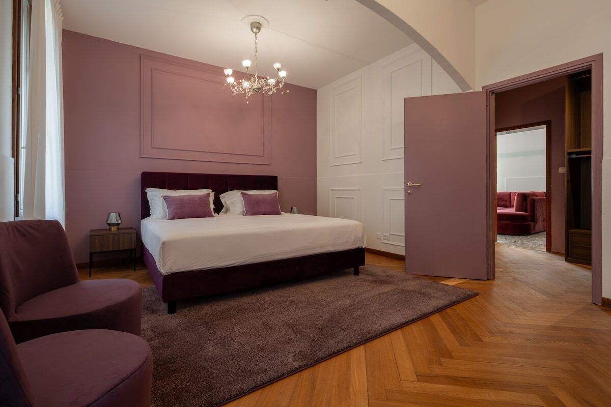 A bedroom with a bed, a nightstand, a chair, a chandelier, and a door leading to another room.