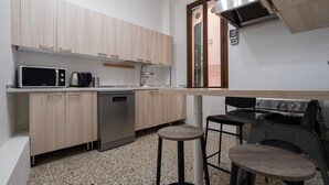 Uma cozinha compacta equipada com micro-ondas, máquina de café, lava-louças e fogão a gás. Há dois taburetes e uma mesa pequena.