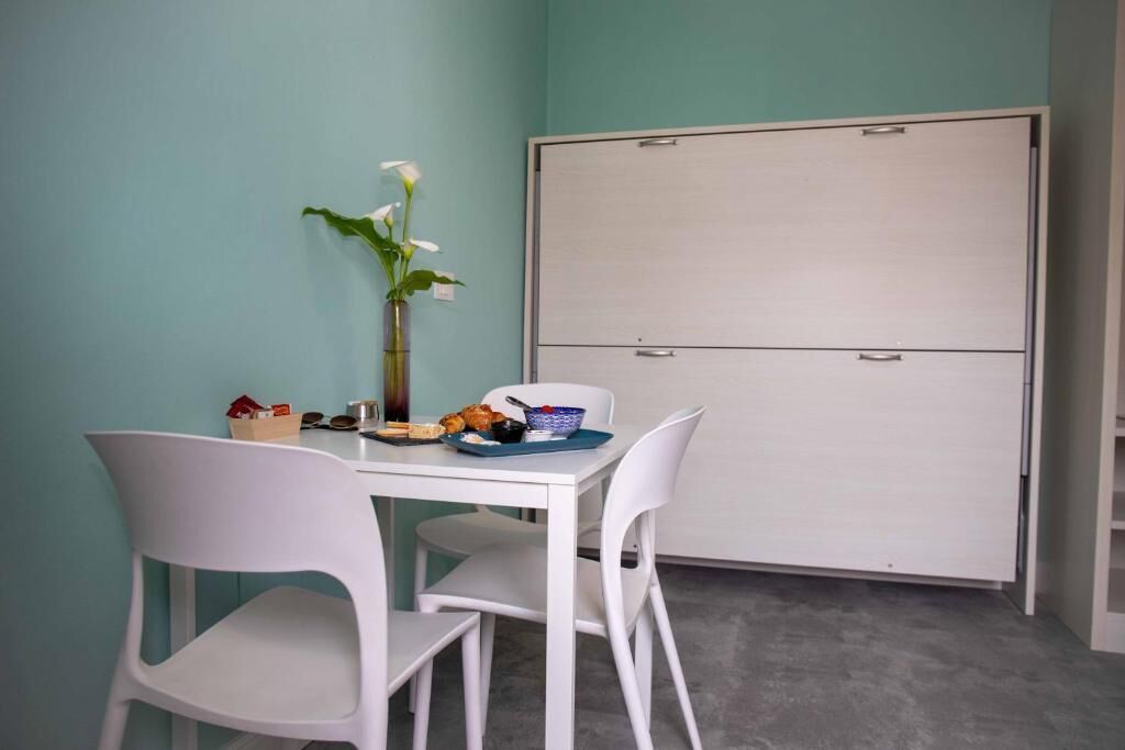 Una habitación con una mesa blanca, dos sillas blancas, un florero con flores, una pizarra blanca y un armario.