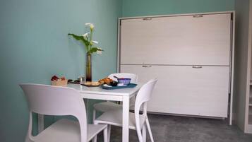 Una habitación con una mesa blanca, dos sillas blancas, un florero con flores, una pizarra blanca y un armario.