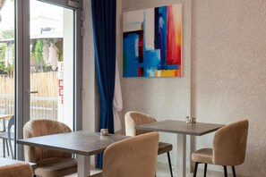 A modern interior with a dining area featuring two tables and chairs, a large abstract painting on the wall, and a view through a glass door.