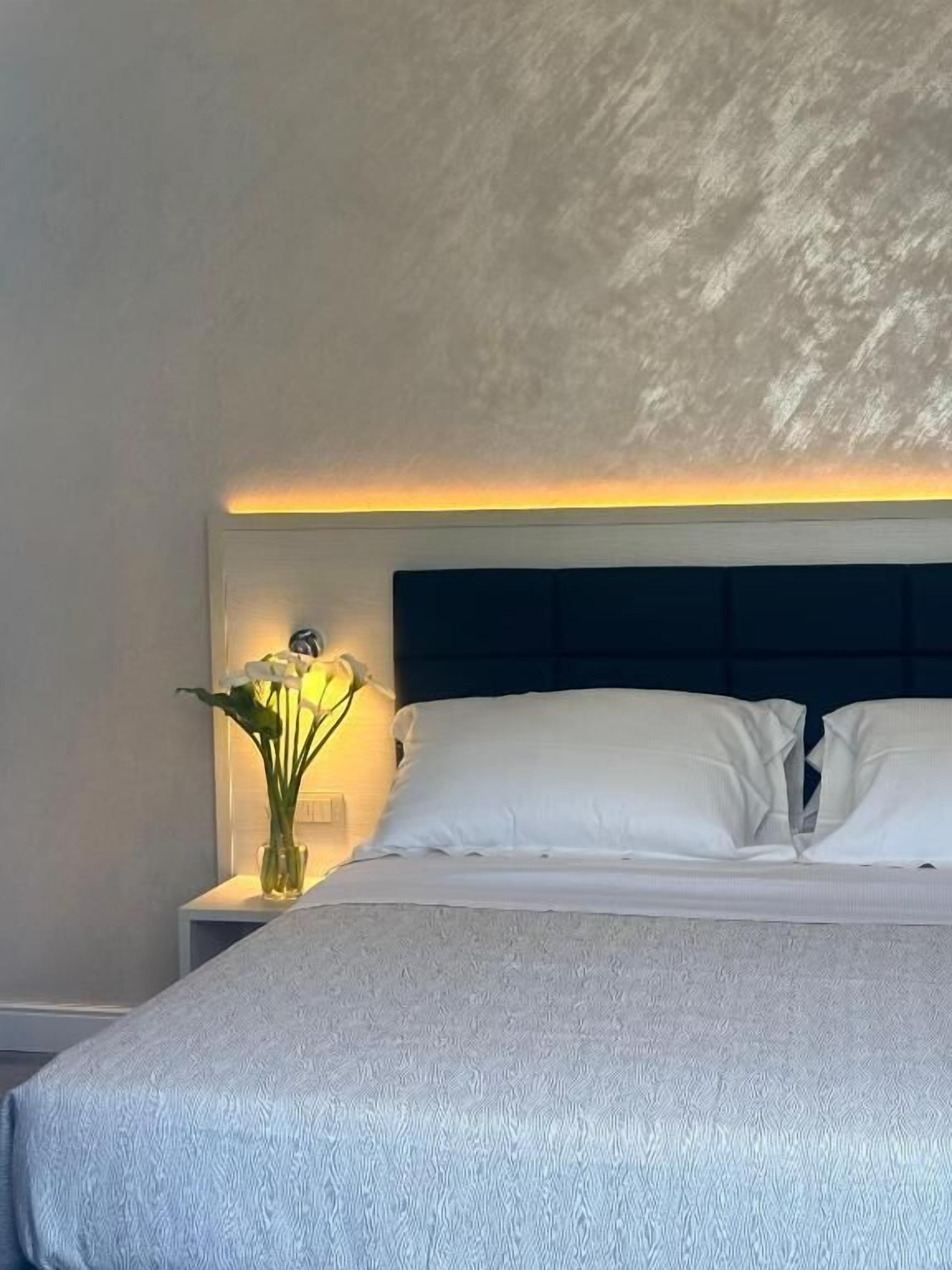 Una cama bien tendida con sábanas blancas, un cabecero oscuro y una mesita de noche con un jarrón de flores.