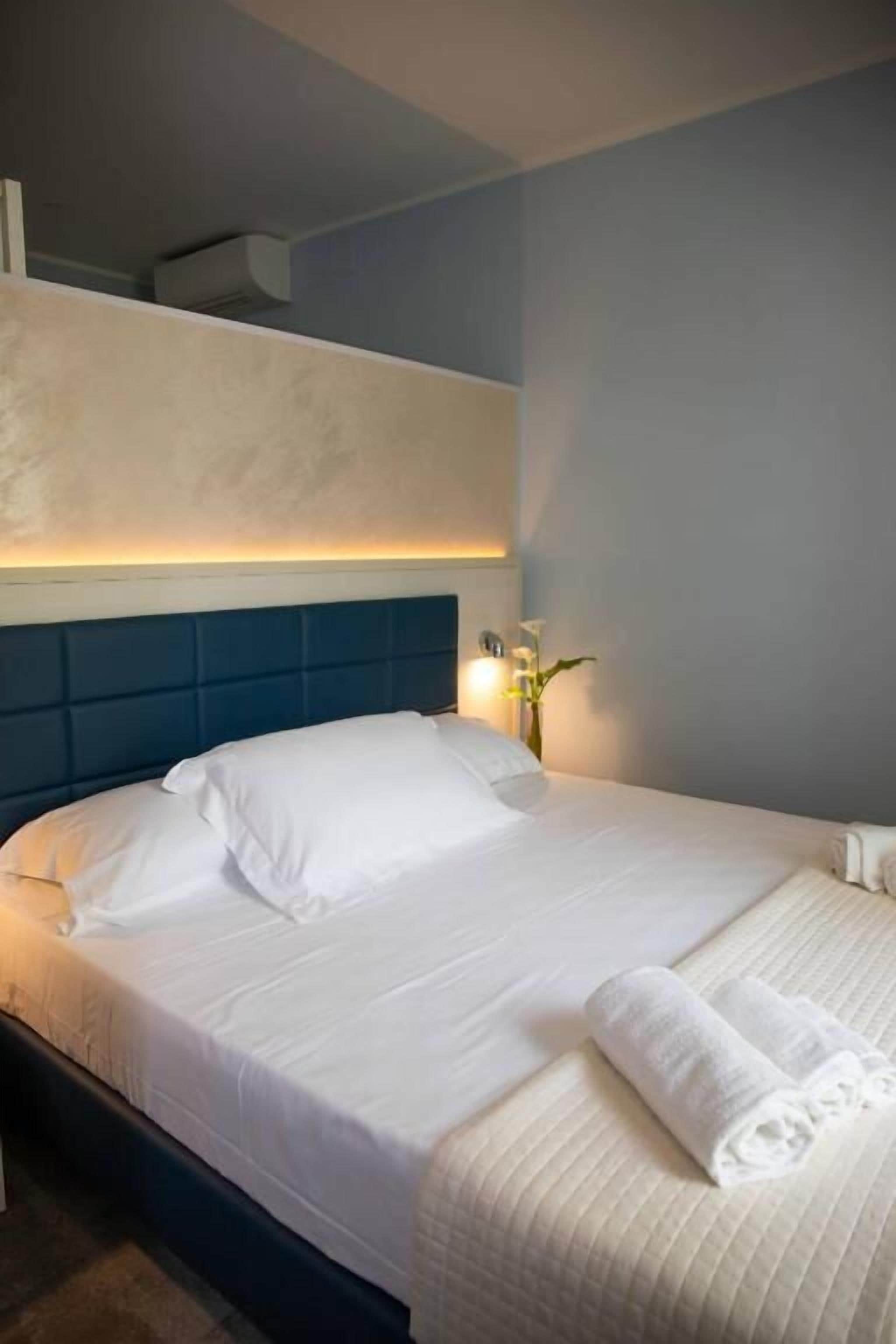 Una cama bien tendida con sábanas blancas, un cabecero azul y una lámpara de noche.