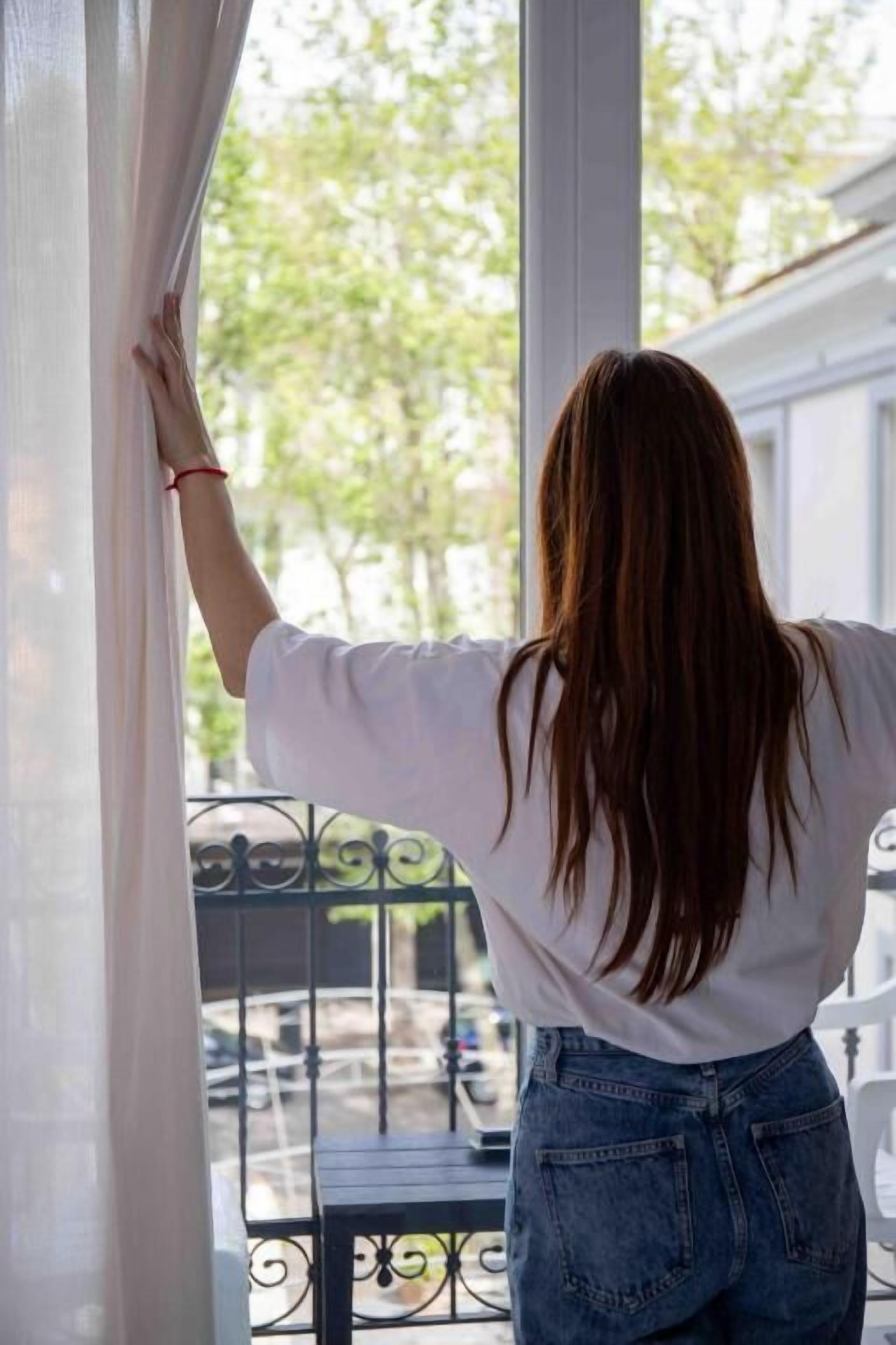Una persona de pie junto a una ventana con vistas a un balcón y mobiliario exterior.