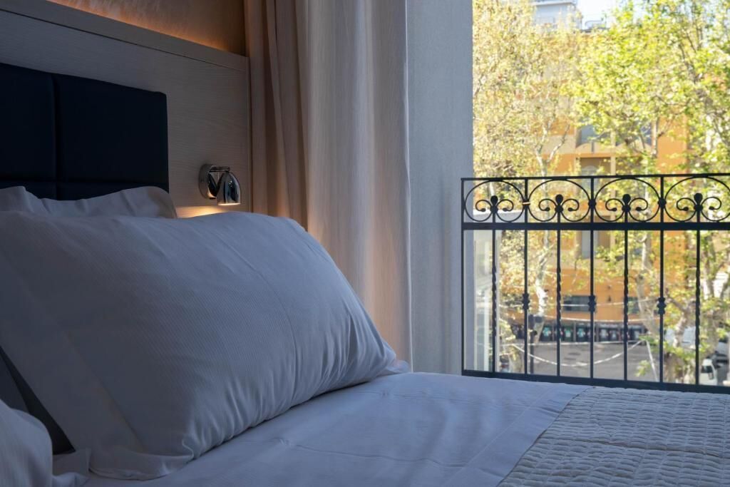Habitación de hotel con una cama, una mesita de noche, una ventana que ofrece vistas a árboles y edificios, y una barandilla de balcón.
