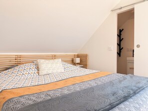 Quarto com teto inclinado, cama com roupa de cama estampada, mesa de cabeceira com abajur e um cabideiro.