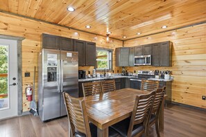 A wooden-paneled kitchen with stainless steel appliances, a dining table with chairs, and a fire extinguisher. - Smoky Ridge Retreat | 2 Cabins, Views & Games (Sevierville)