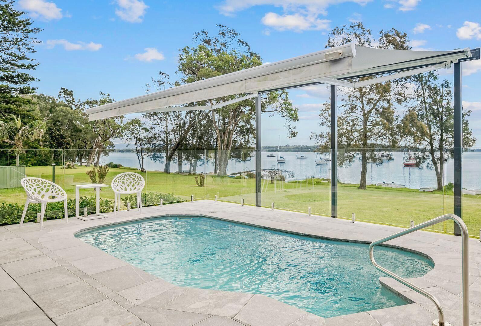 A swimming pool with a glass enclosure, surrounded by a patio area with chairs and a table.