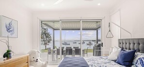 A bedroom with a bed, a desk, a chair, and a view of a body of water through a sliding glass door. - The View | Luxury Lake Macquarie Escape with Pool & Panoramic Water Views (Summerland Point)