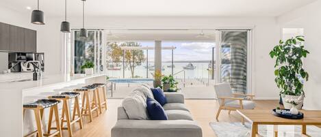 A modern living room with a sofa, a dining table, and bar stools. There is a view of a pool and boats through large windows.