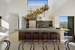 A modern kitchen with a central island, bar stools, and a hanging floral arrangement. - Noa by the Lake Queenstown by MajorDomo | Grand Lakefront Retreat for 14 with Spa Pool, Pilates Studio & Private Gym (Lake Hayes Estate)