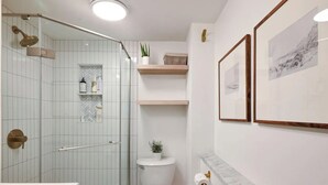 Ein Badezimmer mit Glasdusche, Toilette, Waschbecken und Regalen mit Pflegeprodukten.