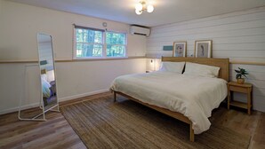 Ein Schlafzimmer mit einem hölzernen Bett, einem Stehspiegel, einem Fenster mit Blick auf Bäume, einem Nachttisch mit Pflanze und einer Wand-Klimaanlage.
