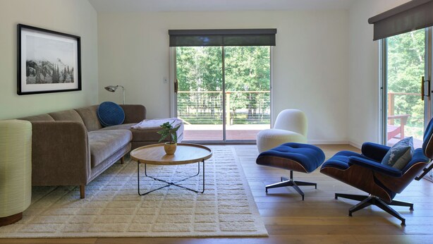 Ein Wohnzimmer mit Sofa, Sessel, Couchtisch und Blick auf einen Balkon mit Grünpflanzen.