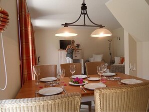 A dining area with a wooden table set for four, surrounded by wicker chairs, and a television in the background. - Gemutliches Ferienhaus mit Terrasse Nahe dem Muritzsee in Rechlin (Rechlin)