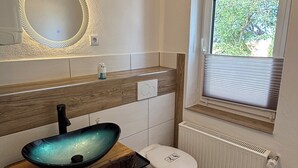 Une salle de bain avec un meuble-lavabo en bois, un miroir rond, des toilettes et une fenêtre avec des stores.