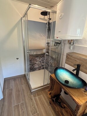 A bathroom with a glass shower enclosure, a wooden vanity with a glass basin, and a white wall-mounted sink. - Vacation Rental: A Short Getaway (Karbach)