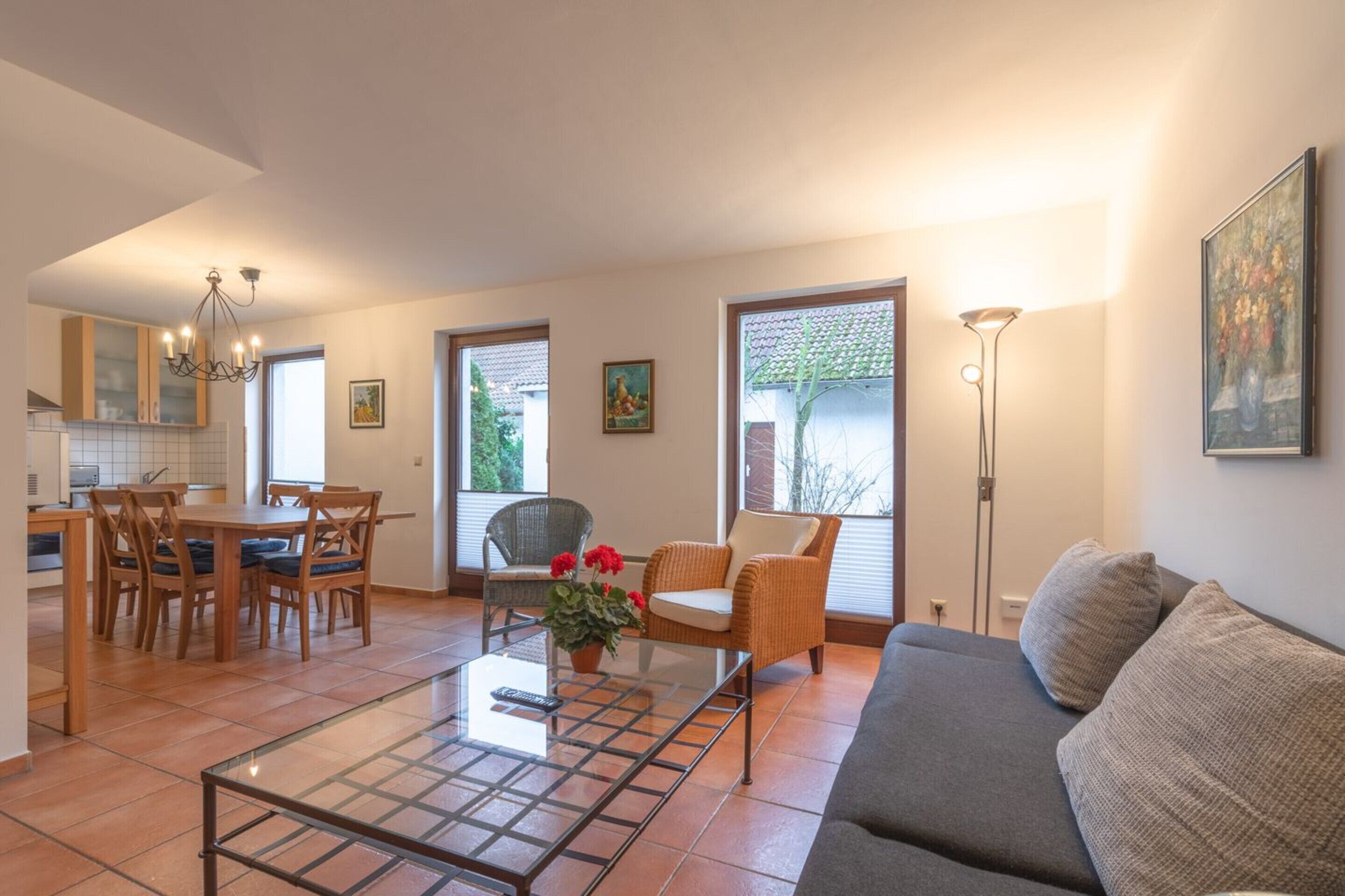 A spacious living area with a dining table, chairs, a sofa, and a coffee table. There are two windows providing natural light and a view of the outdoors.