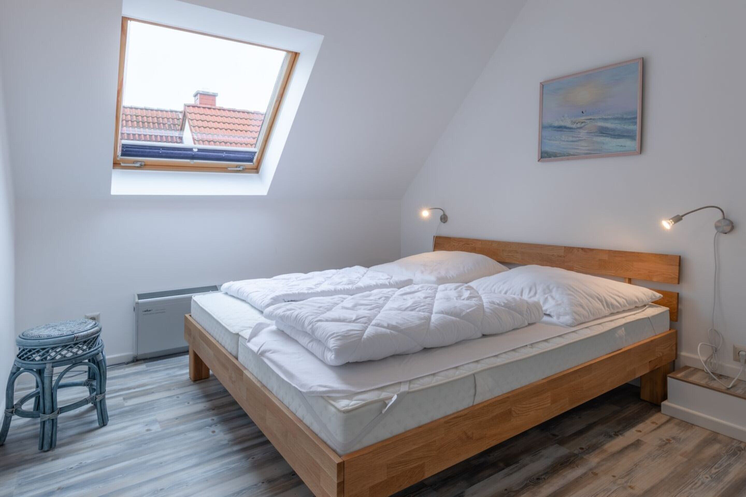 Een slaapkamer met schuine wand, een bed met wit beddengoed, een klein krukje, een verwarming en een schilderij aan de muur.