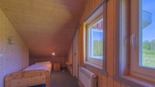 Una cabaña pequeña de un solo ambiente, con techo inclinado, una cama, una mesita de noche, un radiador y una ventana con vista a follaje.