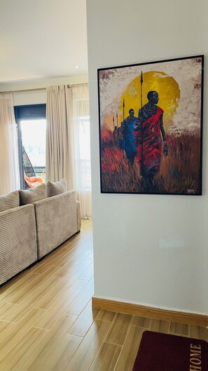A modern living room with a large painting, a sofa, and a view through a window. - Think cozy loft living, open space, and floor-to-ceiling views of Lake Victoria. (Kampala)