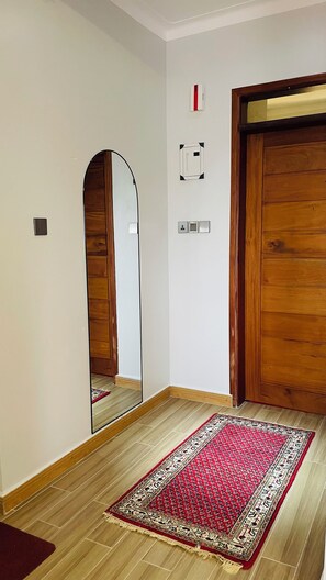 A hallway with a wooden floor, a red patterned rug, a wooden door, a mirror, and a wall-mounted thermostat. - Think cozy loft living, open space, and floor-to-ceiling views of Lake Victoria. (Kampala)