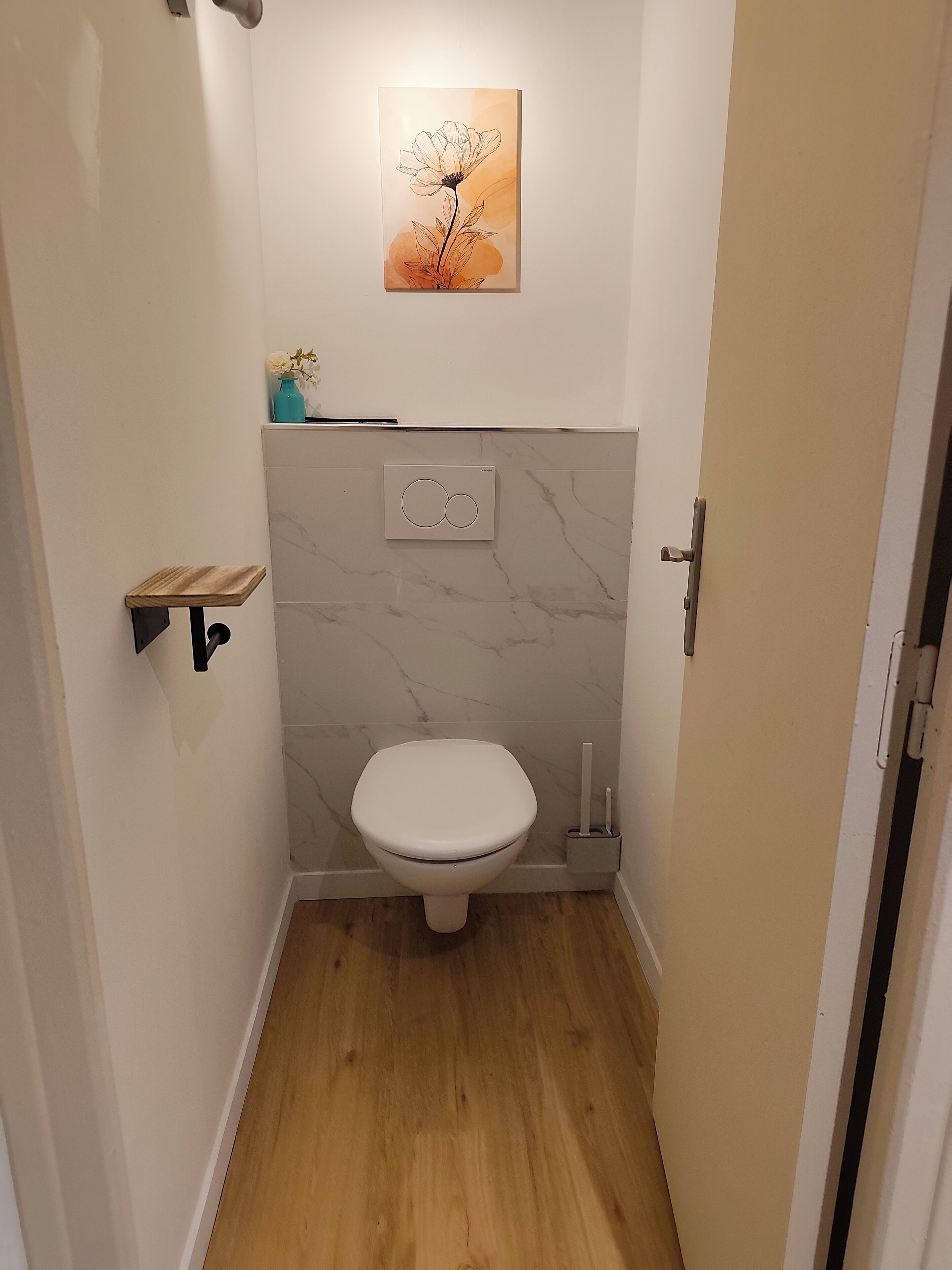 Ein kleines Badezimmer mit Toilette, einer Marmorwand, Holzboden, einer kleinen Ablage mit einer Vase und einem Gemälde über der Toilette.