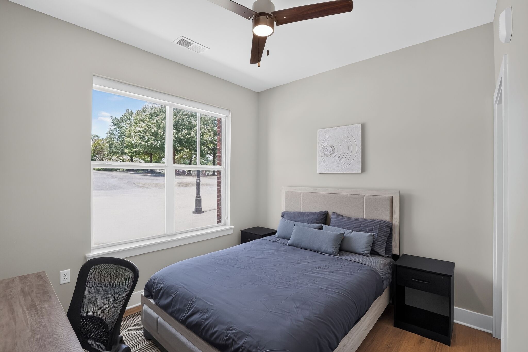 A bedroom with a bed, a desk, a chair, a ceiling fan, and a window with a view of trees.