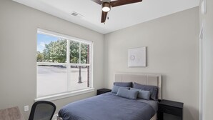 Un dormitorio con cama, escritorio, silla, ventilador de techo y ventana con vista a árboles.
