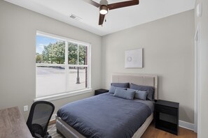 A bedroom with a bed, a desk, a chair, a ceiling fan, and a window with a view of trees.