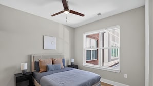 Ein Schlafzimmer mit Bett, Nachttischen, Deckenventilator, Fenster mit Blick und Wandbild.