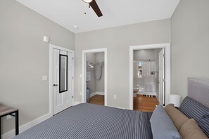 Ein Schlafzimmer mit Bett, Nachttisch, Deckenventilator und Blick ins Badezimmer.
