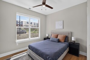 A bedroom with a bed, bedside tables, a ceiling fan, a window, and a view of buildings outside. - Homey 2BR with Great Space (Greer)