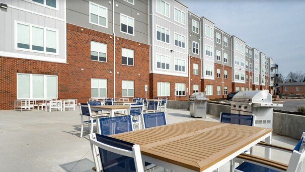 An outdoor patio with tables and chairs, a barbecue grill, and a building with brick and white siding. - Homey 2BR with Great Space (Greer)