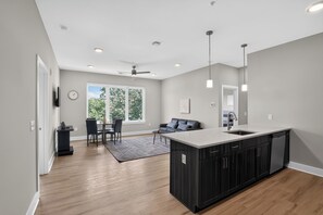 A modern kitchen with a central island, a dining area with a table and chairs, a sofa, and a large window with a view of greenery. - Parkview Greer  2 Bedroom Haven (Greer)