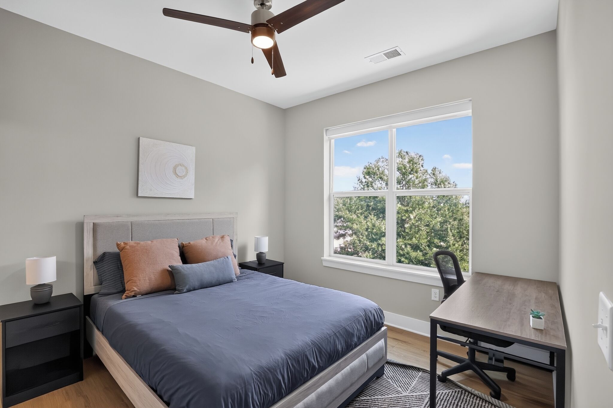 A bedroom with a bed, nightstands, a desk, a chair, a ceiling fan, and a window with a view of trees.