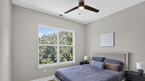 Ein Schlafzimmer mit einem Bett, einem Deckenventilator, einem Fenster mit Blick auf Bäume und einem Nachttisch mit einer Lampe.