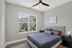 A bedroom with a bed, a ceiling fan, a window with a view of trees, and a bedside table with a lamp. - Parkview Greer  2 Bedroom Haven (Greer)