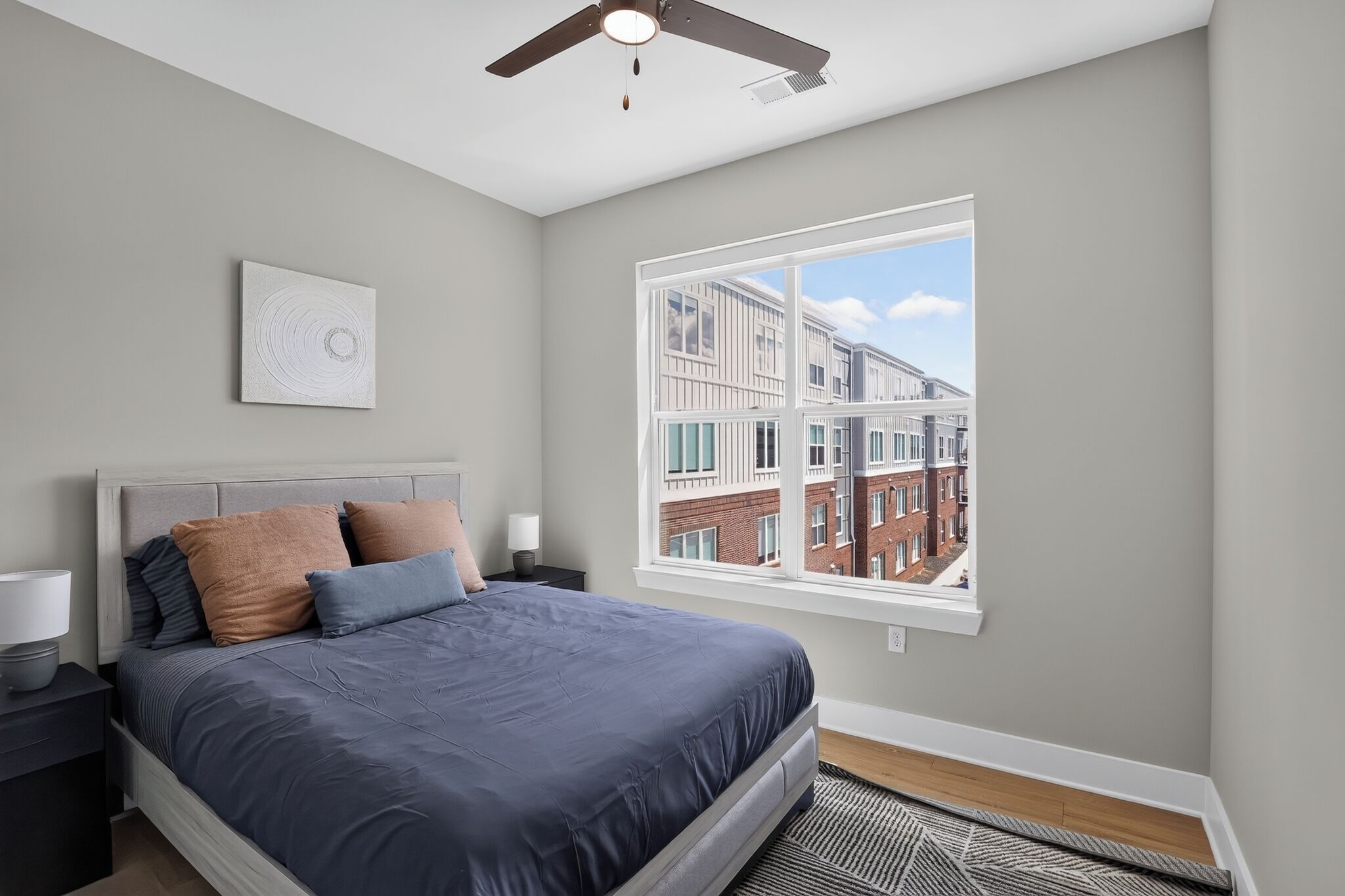 A bedroom with a bed, a ceiling fan, a window with a city view, and a wall-mounted artwork.