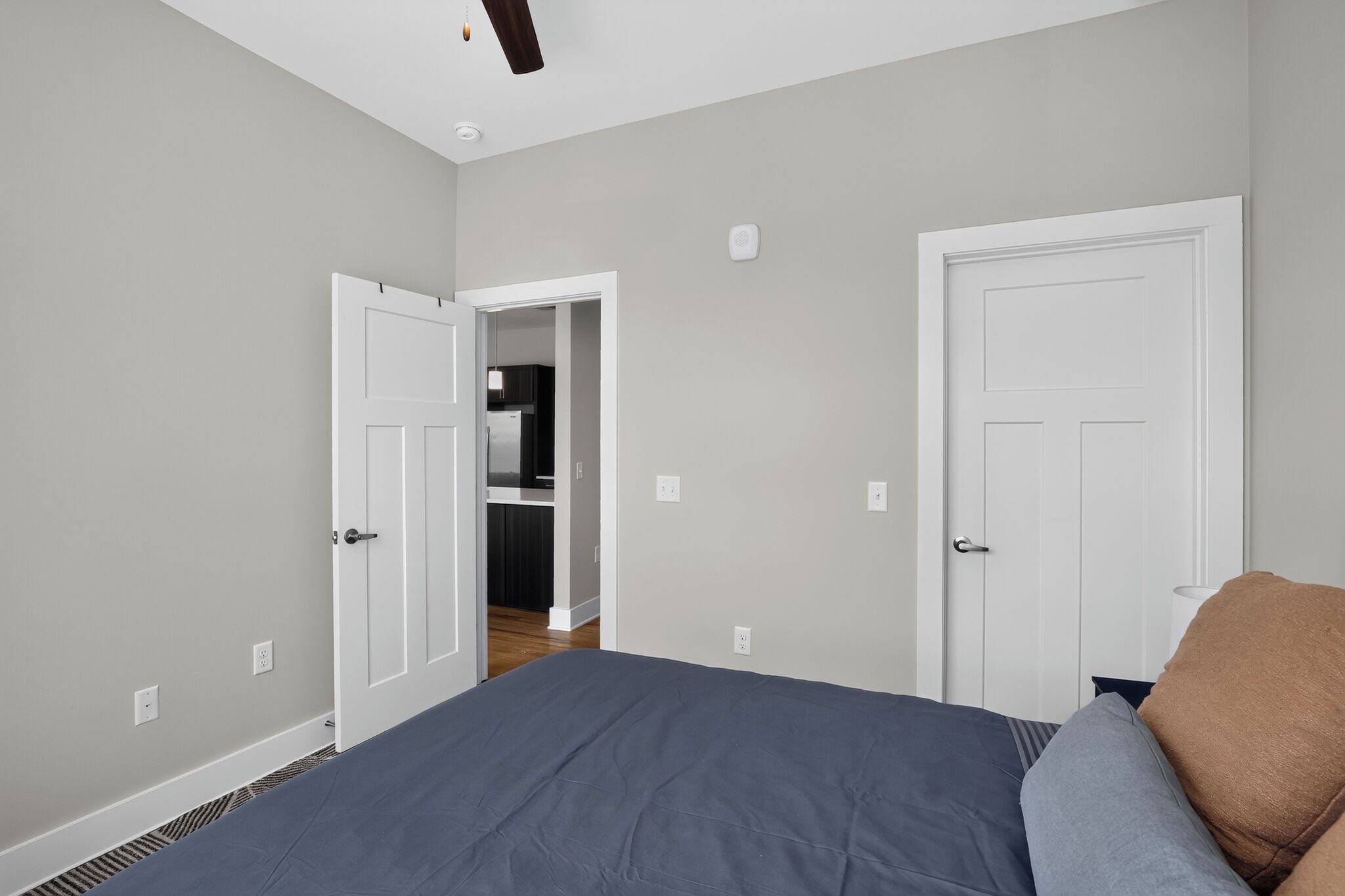 A bedroom with a bed, two doors, a kitchen in the background, and a ceiling light.