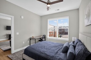 Ein Schlafzimmer mit einem Bett, einem Schreibtisch, einem Stuhl, einem Deckenventilator, einem Fenster mit Stadtblick und einer Tür zu einem weiteren Raum.