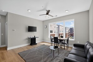 Ein modernes Wohnzimmer mit Flachbildfernseher, Esstisch mit Stühlen, schwarzem Ledersofa, gemustertem Teppich und Deckenventilator.